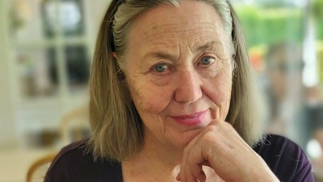 An elderly woman sits at the table, holding her chin with her left hand.