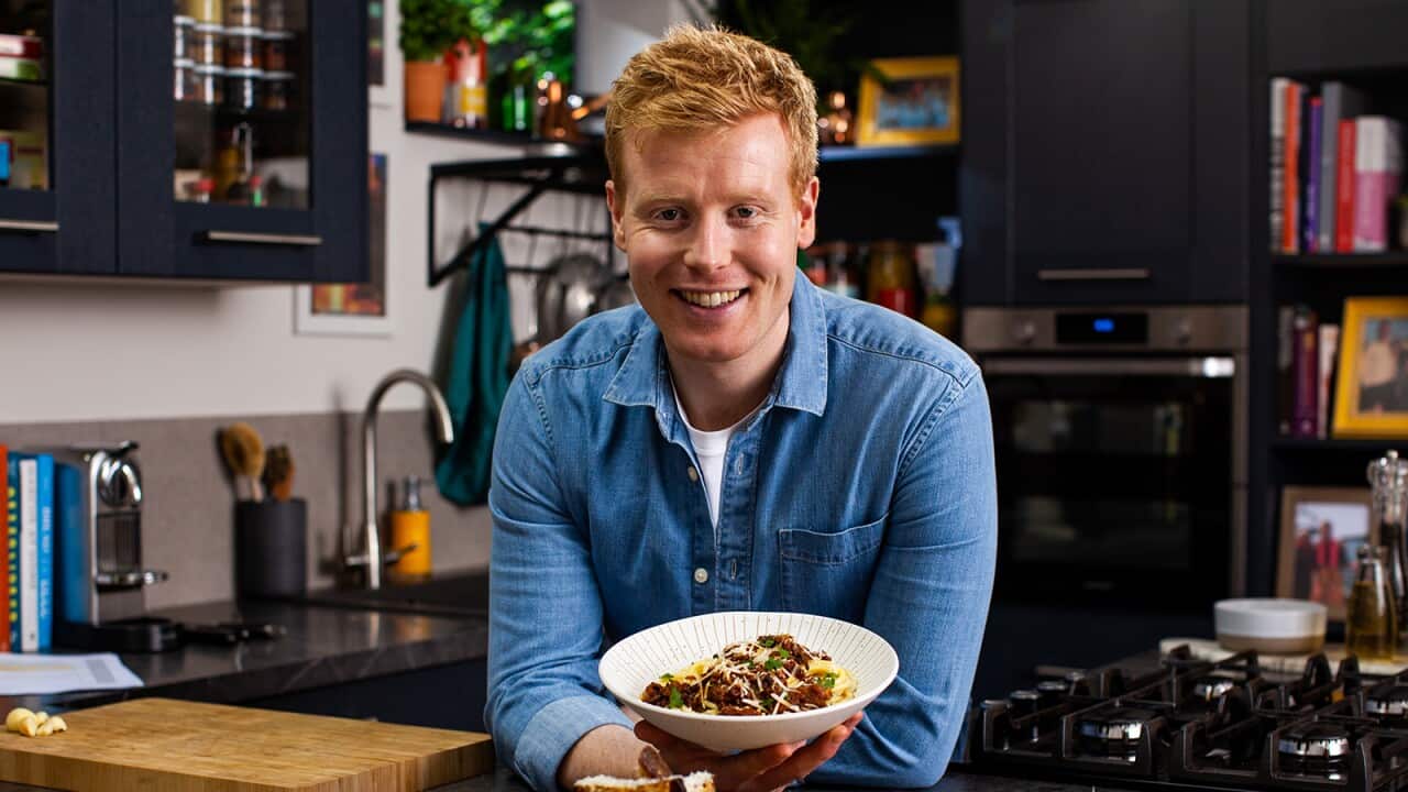 A smiling red-headed man in a blue shirt stands in a kitchen, leaning forward onto a bench with a bowl of food in one hand.