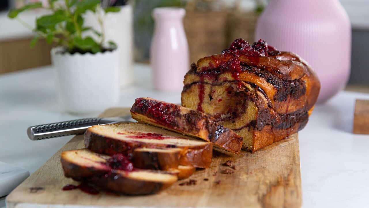 A loaf-shaped babka sits on a wooden board. Three slices have been cut from one end and sit in front of the rest of the loaf. Chocolate-berry swirls run through the loaf.