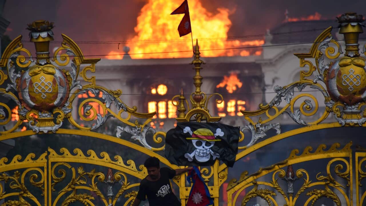 A large building is engulfed in flames behind a golden gate, with a man climbing the gate's bars. He is holding a Nepali flag and a pirate flag featuring a skull with a straw hat is hanging from the gate.
