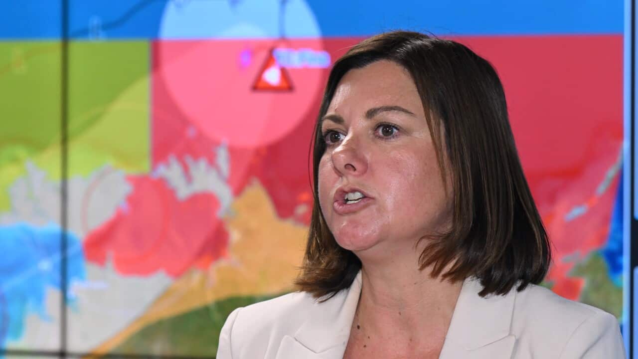 A woman with shoulder-length brown hair and a white blazer speaking in front of a meteorological map.