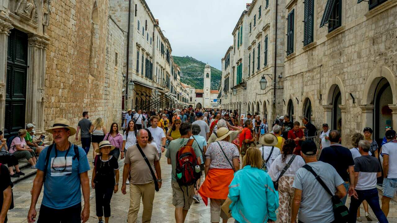 Crowds of people walk down a street fringed by historic stone buildings.