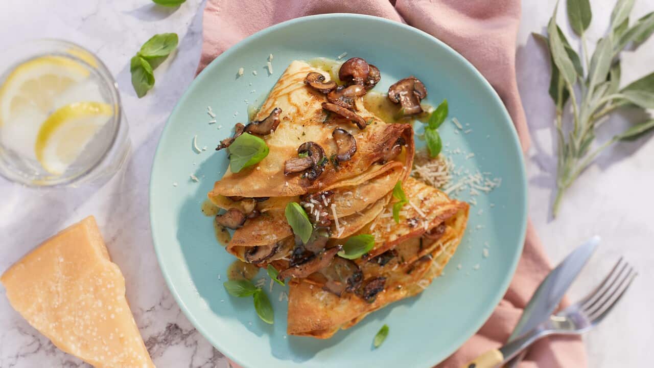 Thin pancakes, folded into quarters and scattered with cooked mushrooms, sit on a blue plate. The plate sits on a pinkish napkin on a marble surface, with bone-handed knife and fork sitting alongside.