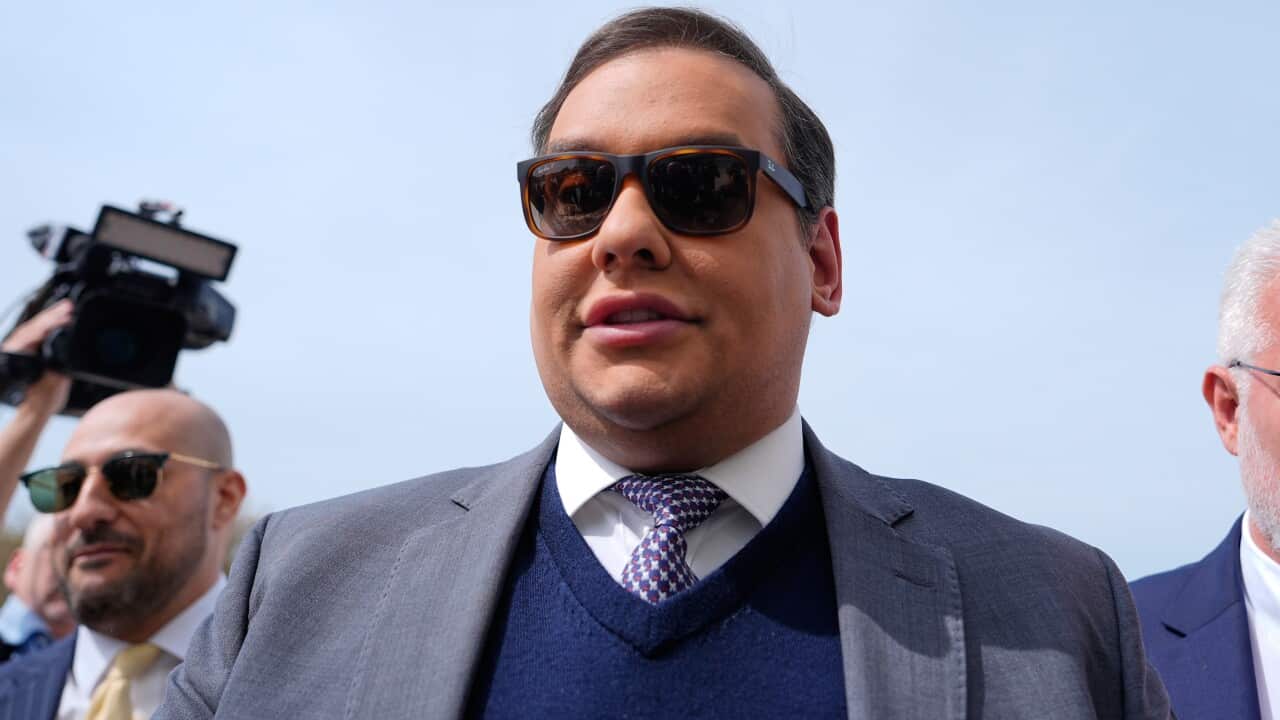 A white man wearing sunglasses, a grey suit jacket, blue sweater and tie.