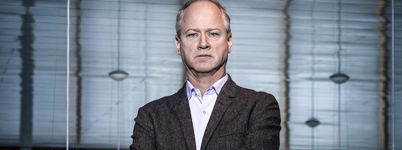 A man in a charcoal suit stands with folded arms, staring straight ahead.