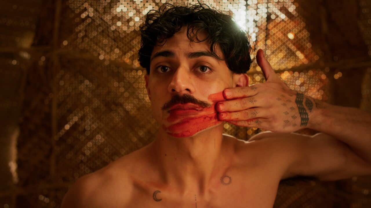 Gamilaroi and Tongan man, Daniel Mateo stands in front of a gold beaded background and wipes his hand across his face, leaving a smear of read paint.