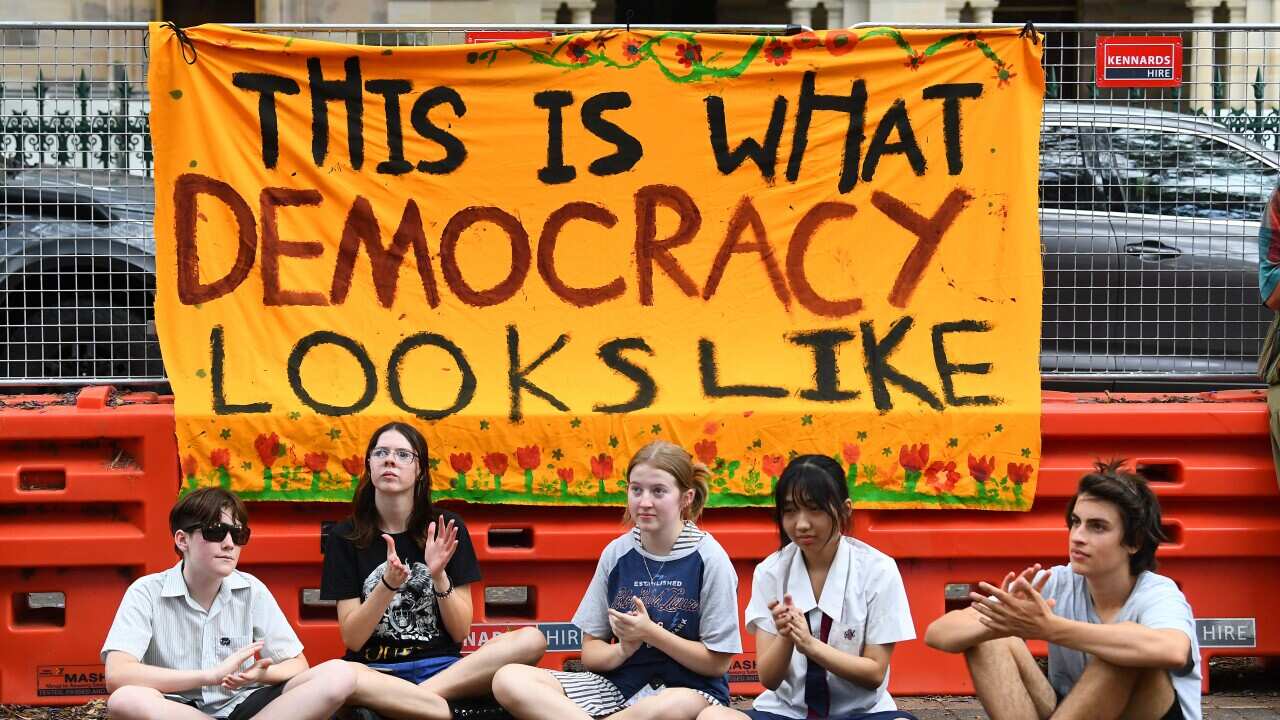 Five teenagers are sitting on the ground under a banner that reads “This is what democracy looks like“ next to a construction site.