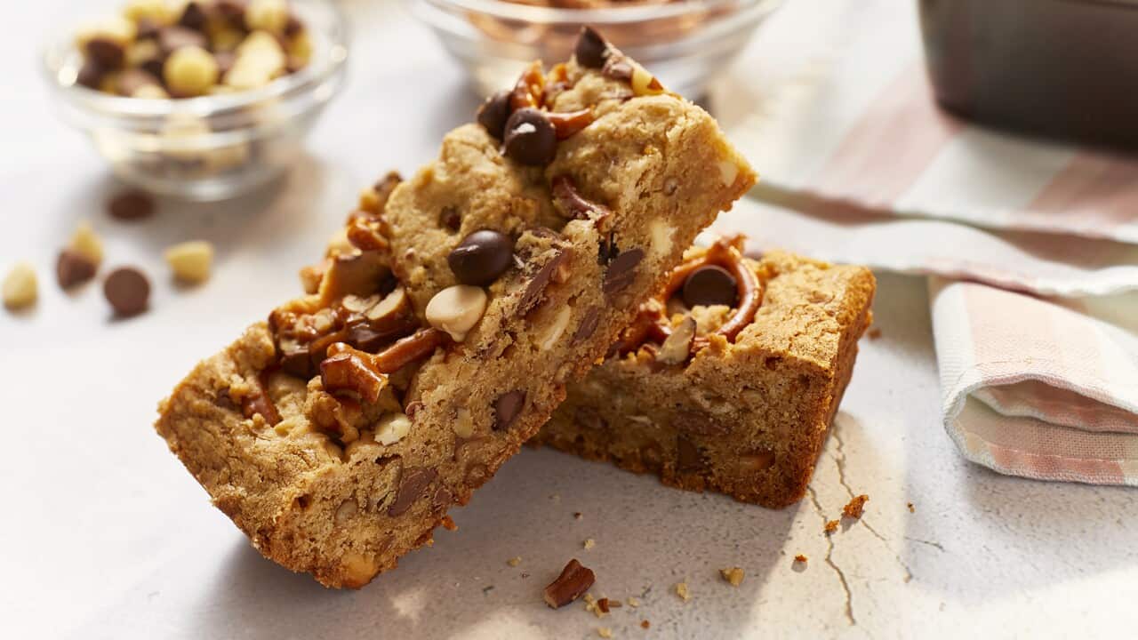 Two rectangular biscuit-like bars sit on a pale surface, one leaning on the other. They are studded with nuts, choc chips and mini pretzels. Two bowls with chocolate chips and pretzels can be seen, out of focus, in the background. A folded pink and white napkin sits alongside the two bars. The edge of a baking container can be seen on the napkin.