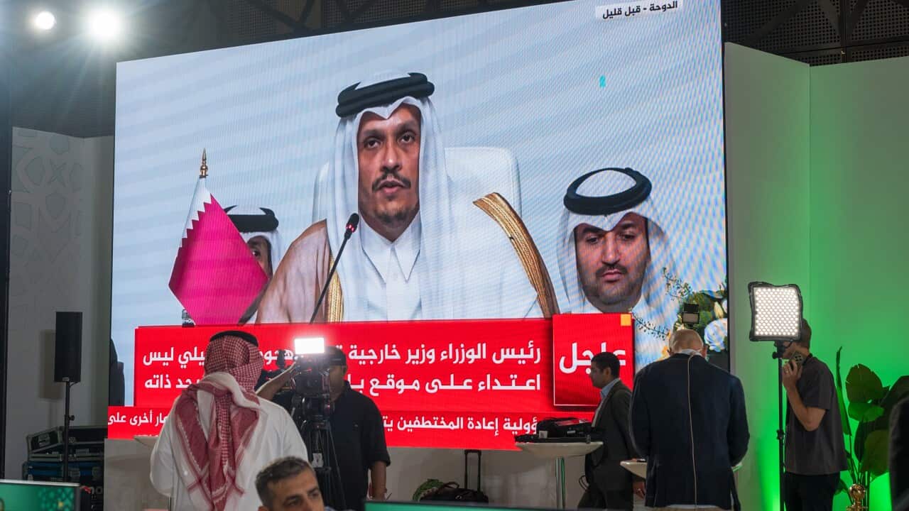 A man is shown on a screen speaking into a microphone. There are various people near the screen watching.