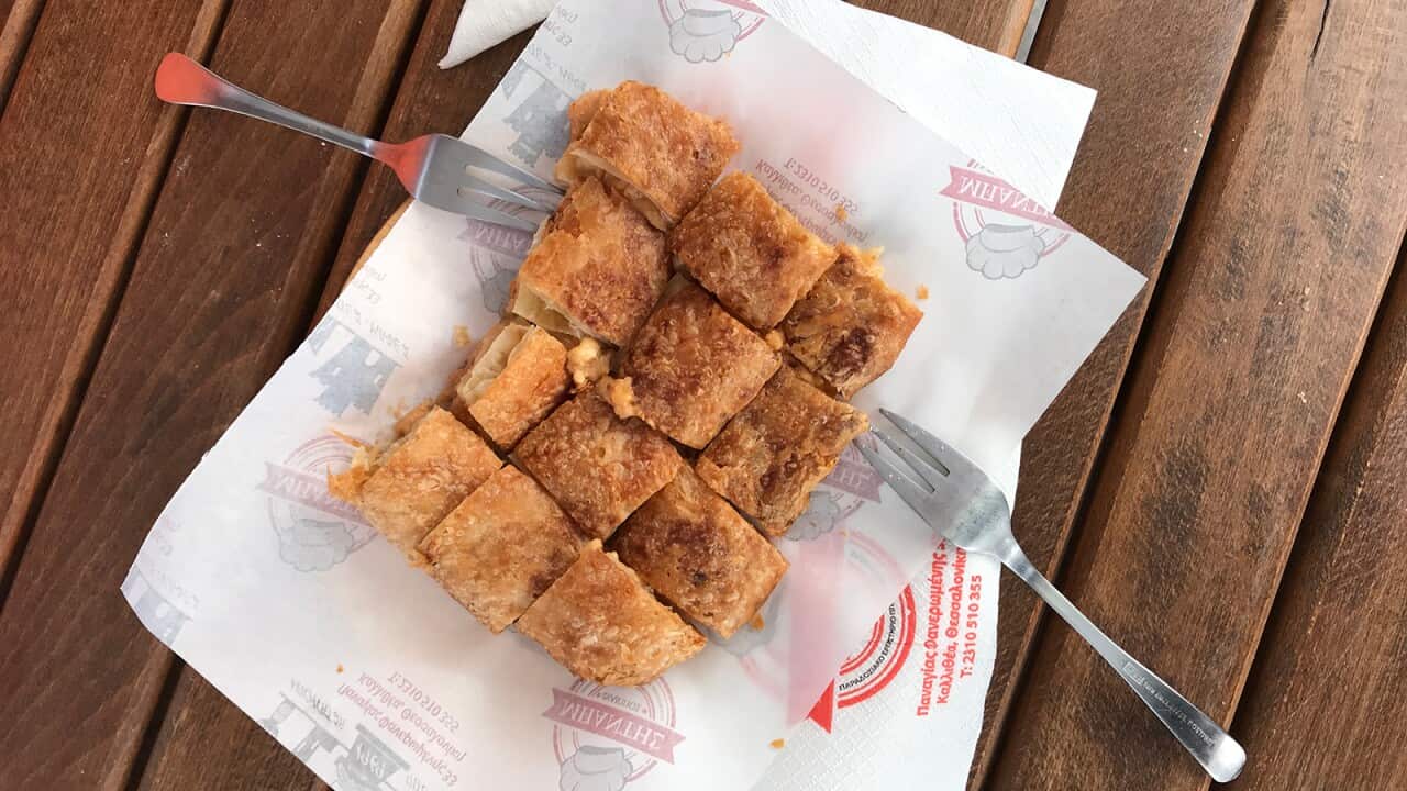 A rectangular-ish block of golden Greek pastry, cut into 12 pieces, sits on two deli-style sheets of paper on a wooden surface. Two forks sit touching the tastry. Custard filling can be seen near two of the pastry squares.