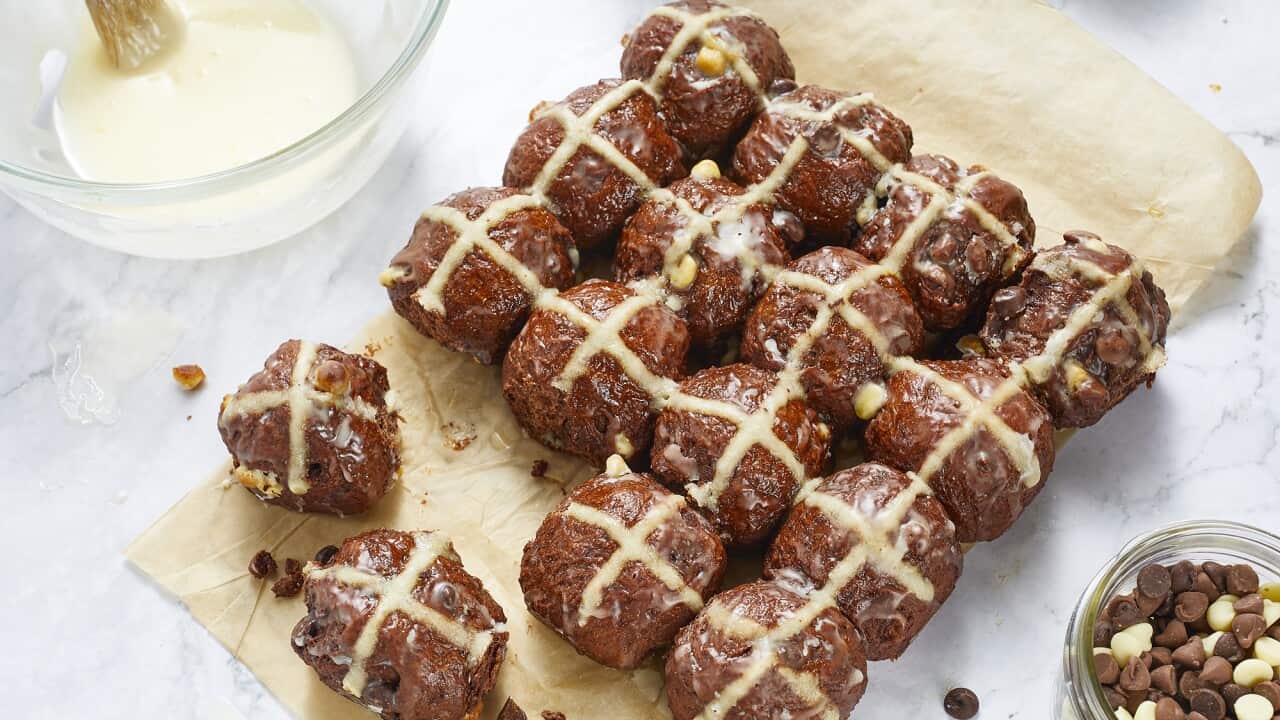 Seen from overhead, a 4x4 array of baked chocolate hot cross buns sits on a piece of baking paper on a marble surface. Two buns have been pulled off the main grouping, and sit slightly apart. A glass jar with white, dark and milk choc chips sits on one side, with a few chips scattered on the marble surface.