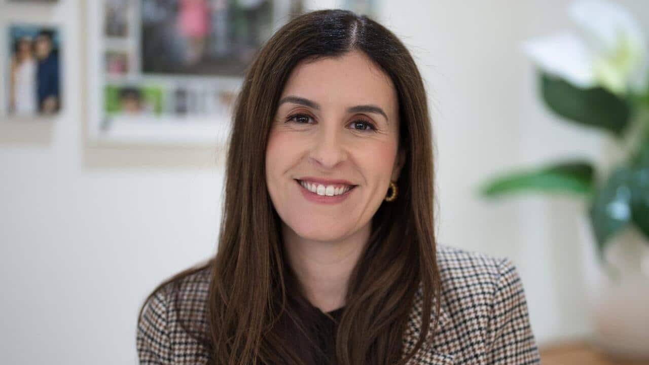 A woman with long brown hair and wearing a checkered blazer is smiling.