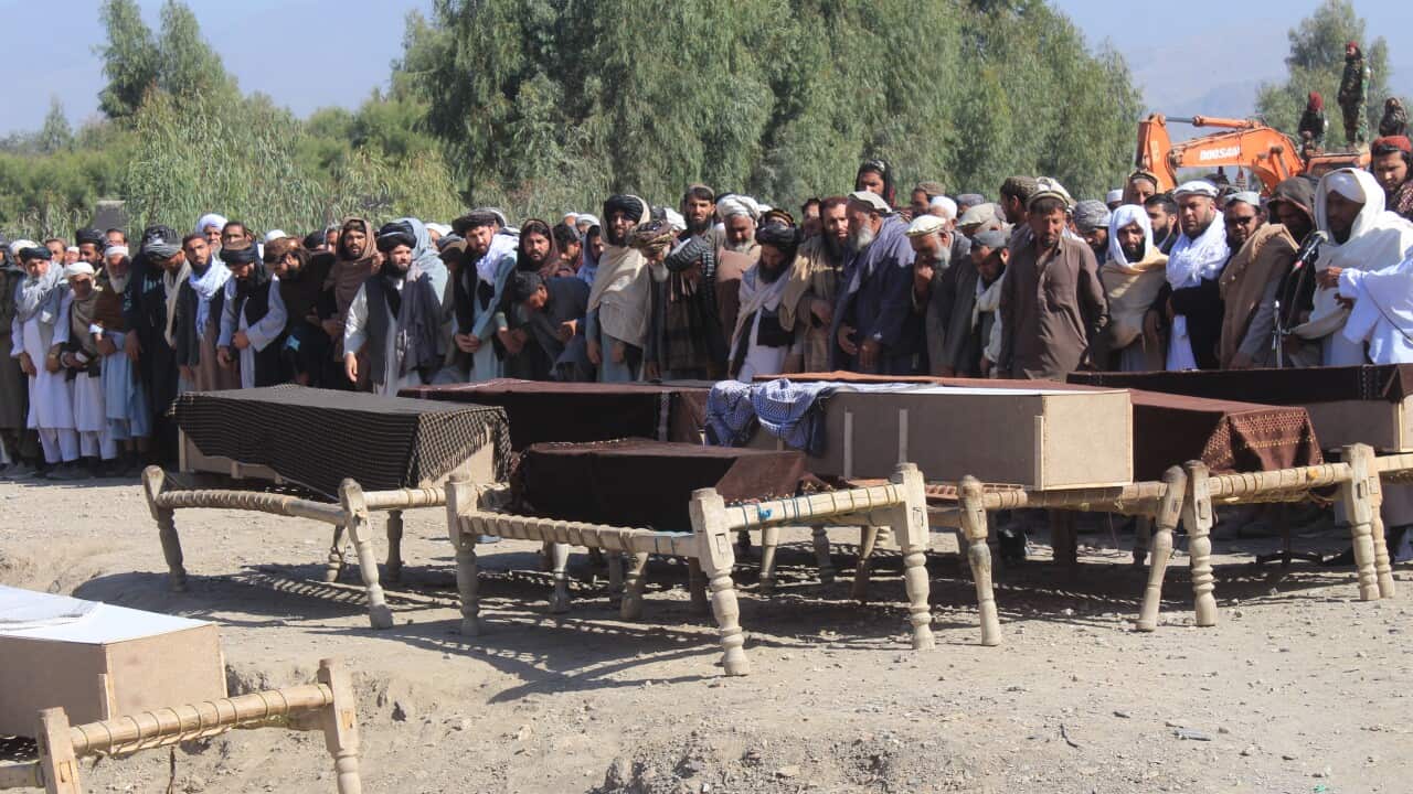 A large group of men stand in prayer behind several coffins laid on wooden biers at an outdoor funeral, with trees and construction equipment in the background.
