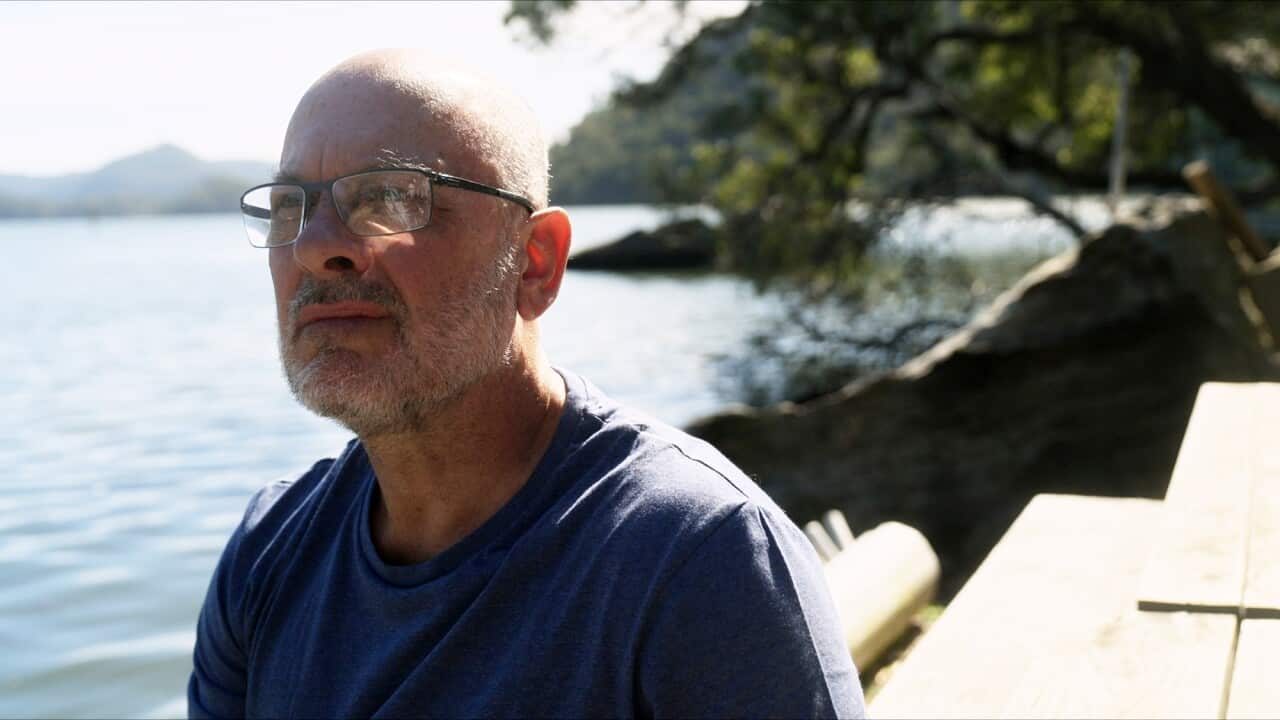 A man in glasses and a blue shirt sits beside a river.