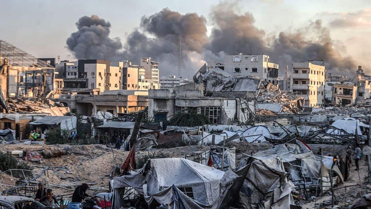 Smoke in the distance rises over damaged and destroyed buildings, tents and people can be seen in the foreground.