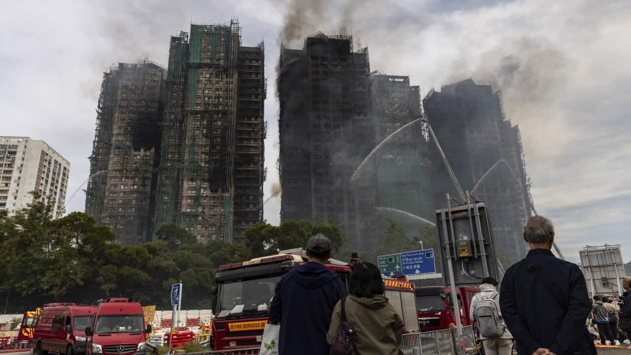People on the street gaze up at multiple severely fire-damaged buildings as smoke rises