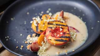 A wide blue-black bowl is half-filled with a creamy custard. Grilled and fresh peaches sit in and beside the custard, garnished with chopped nuts, small white flowers and sliced cumquat.