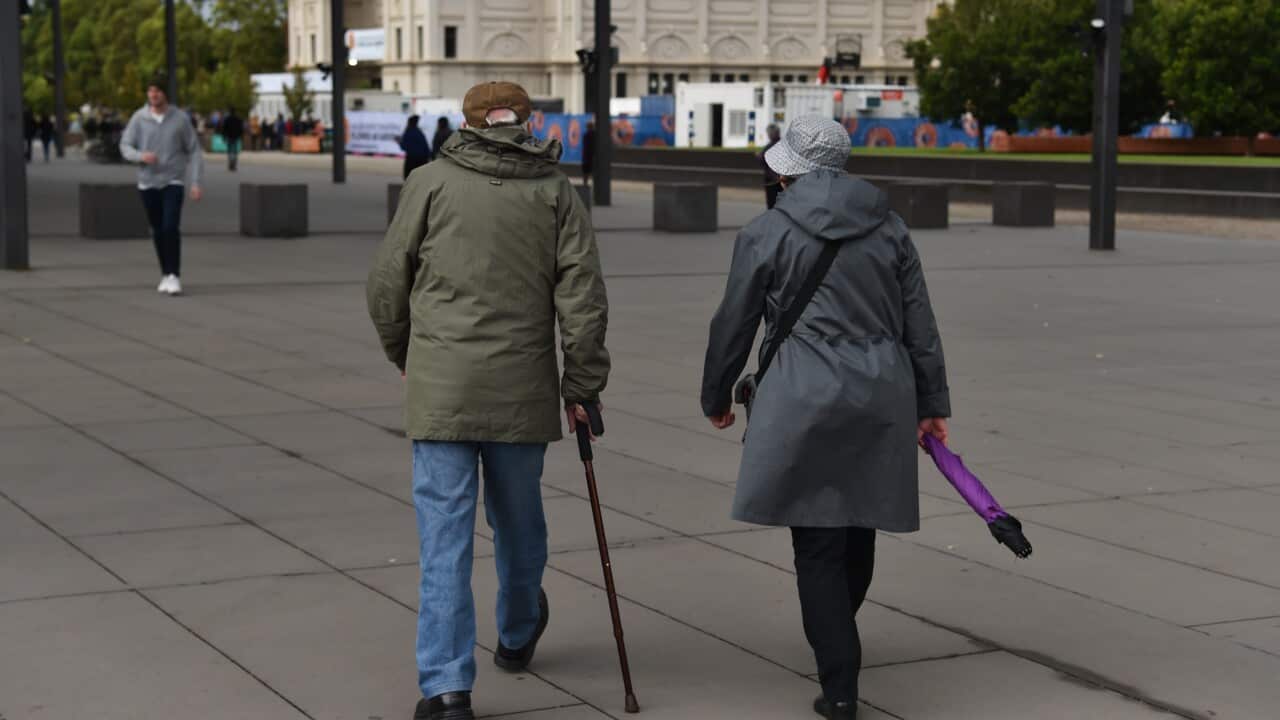 Two elderly people in coats walk away from the camera across a wide paved plaza, one using a cane and the other holding a purple umbrella, with a large ornate building in the background.