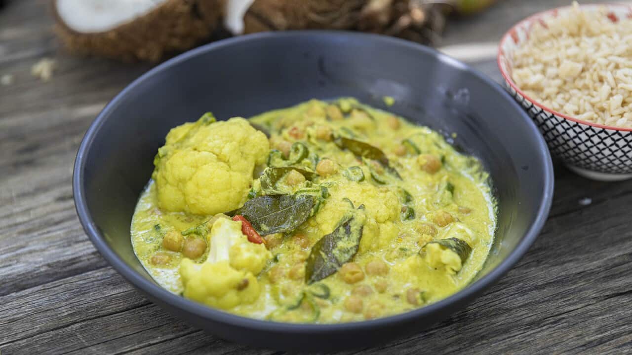 A vibrant yellow curry sits in a dark bowl. A smaller bowl, with rice, sits behind it. An opened coconut can be seen in the background.