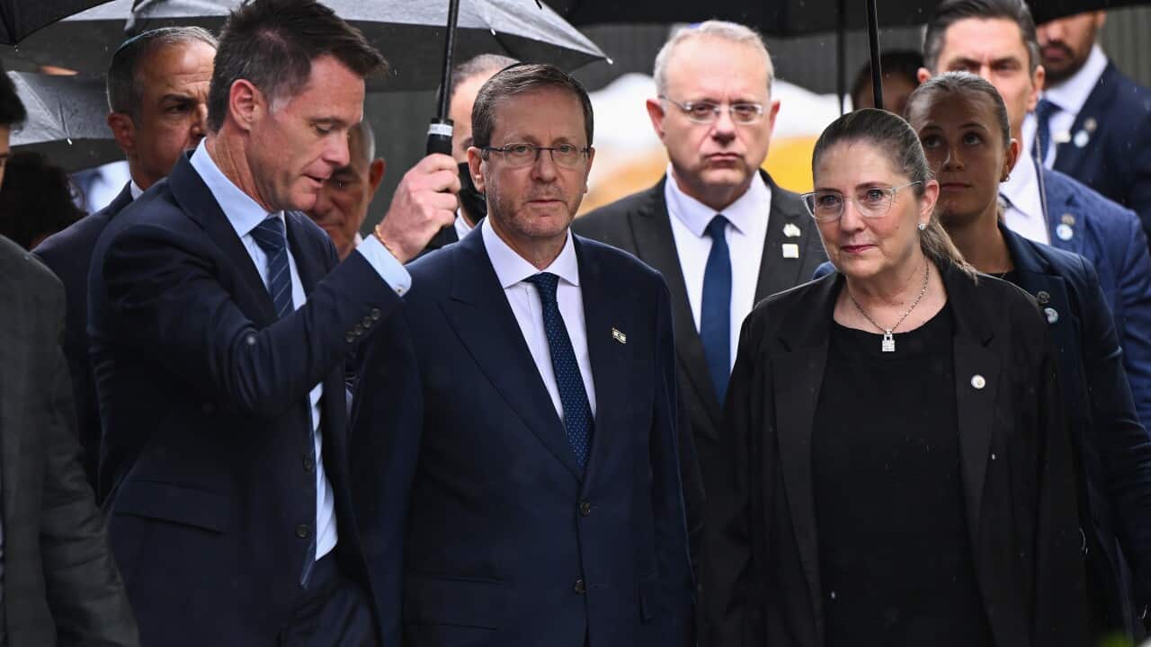 A middle-aged white man in a suit holding an umbrella, a white man and woman stand beside him. Several people are behind them.