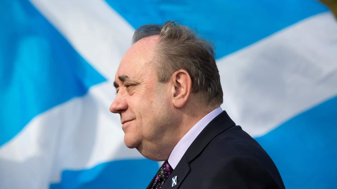 An older man stands side-on in front of a Scottish flag