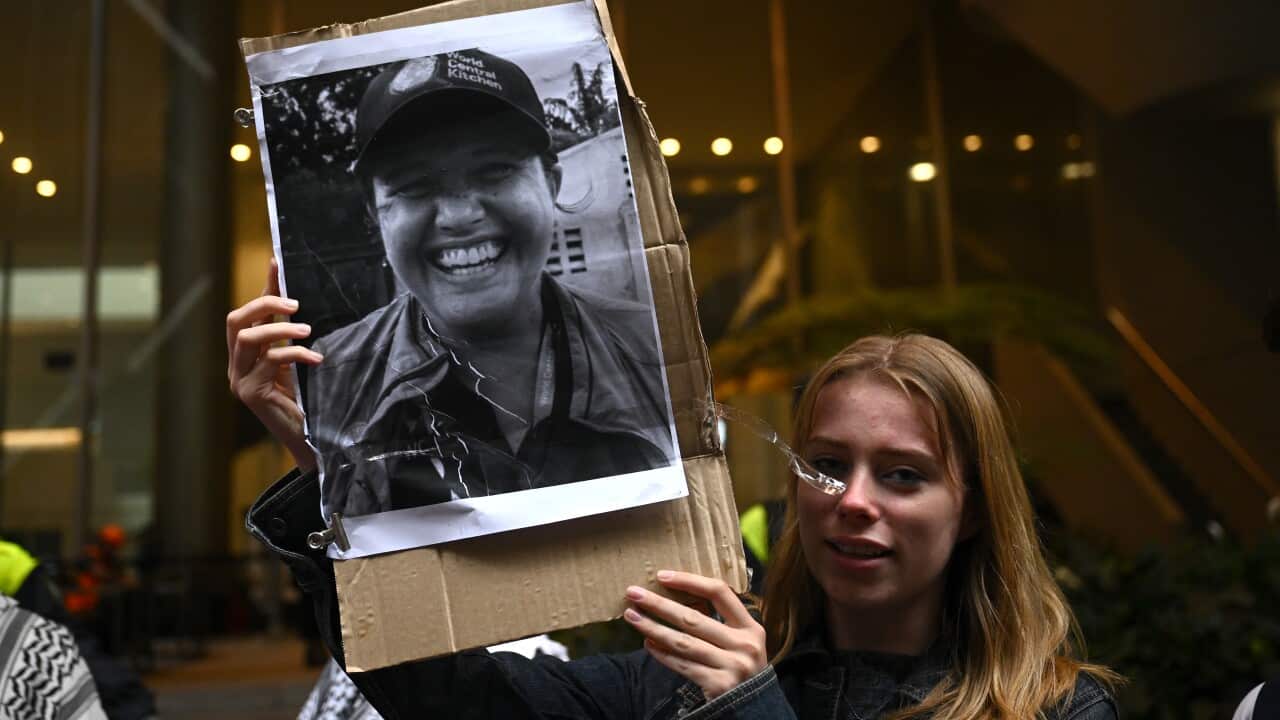 A woman holds up a photo of another woman at what appears to be a protest. Several people wearing Palestinian keffiyeh are visible in the background.