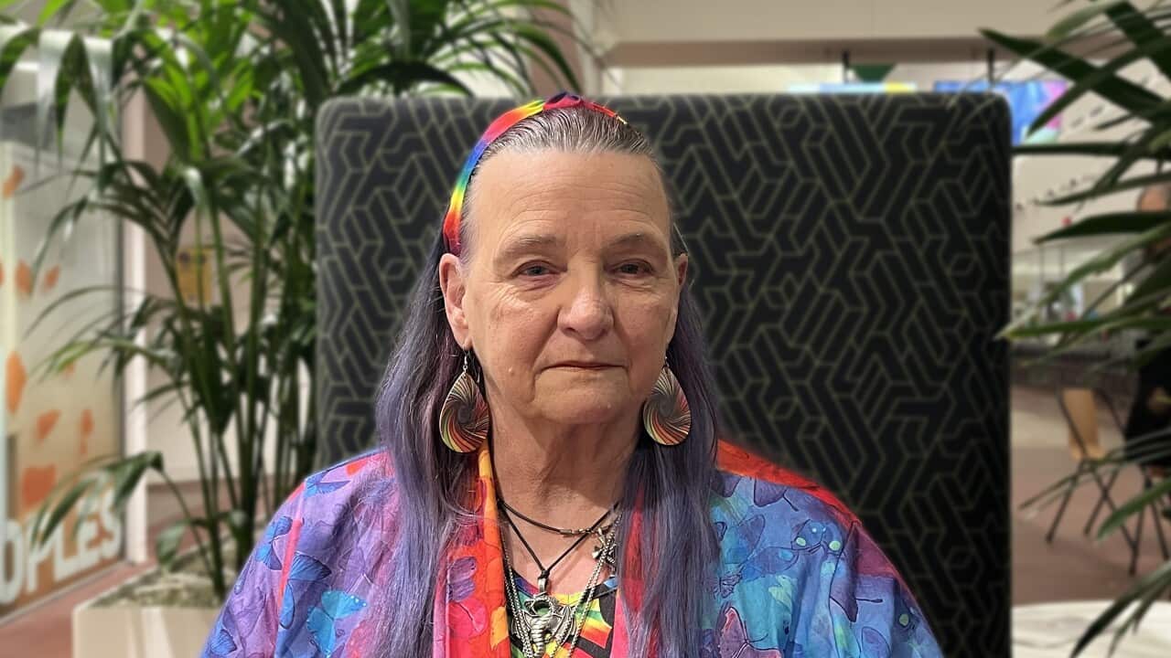 an older woman with long purple hair wearing a rainbow headband, jewellery and clothes looks neutrally at the camera in a closeup in an office