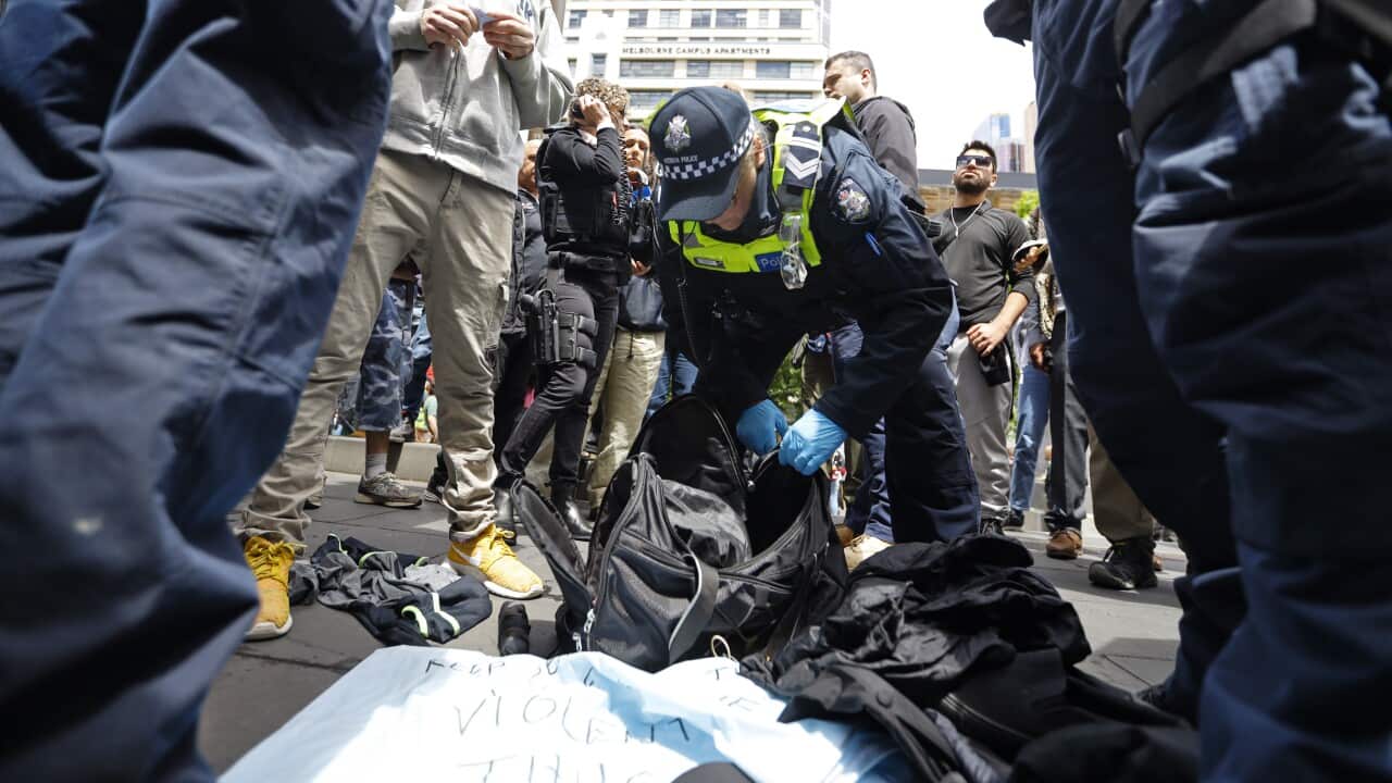 A police officer in a dark blue uniform and gloves searches a black bag on the ground, surrounded by several people in a busy, urban setting.