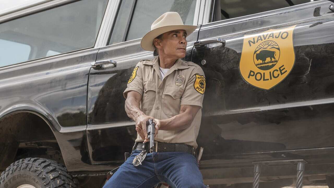 A Native American policeman crouches beside a police vehicle, gun in hand.