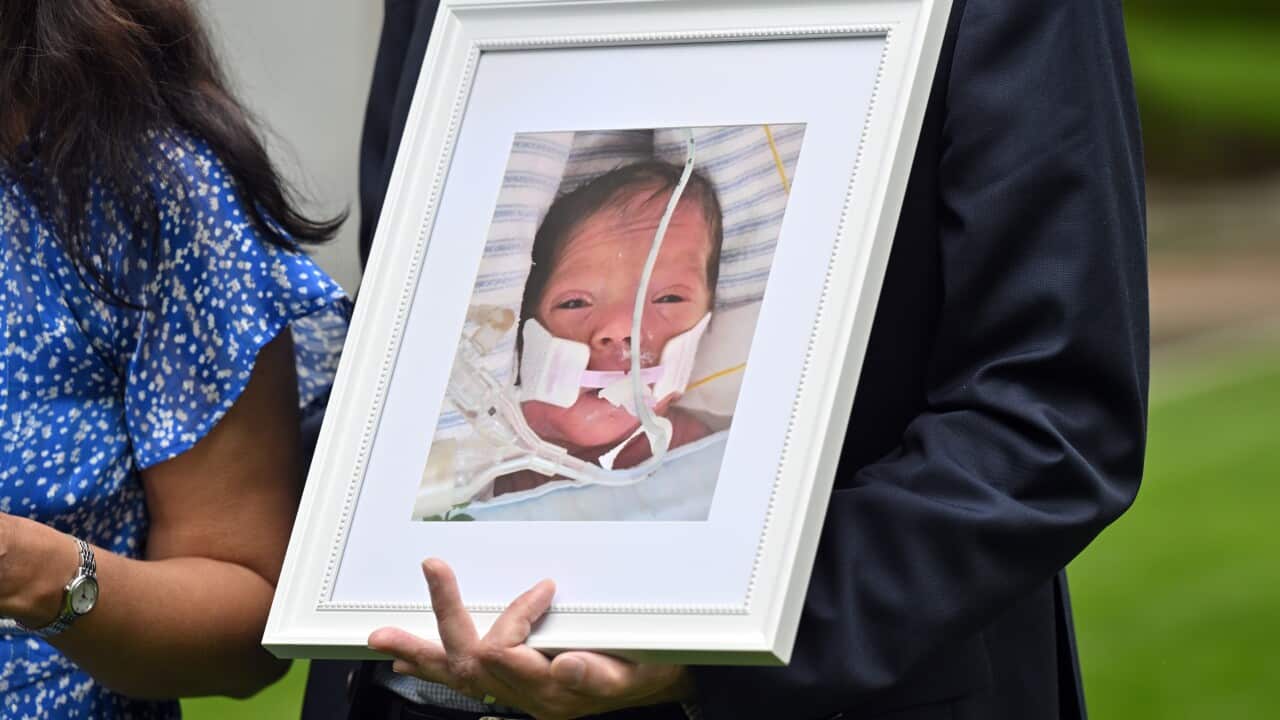 A man wearing a suit holds a white framed photograph of a baby. The baby has a medical tube in her mouth and is lying in a bed with a blanket. The man's head is not visible.