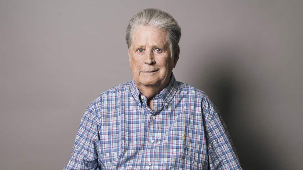 A man with grey hair wearing a blue checked shirt, smiling faintly.