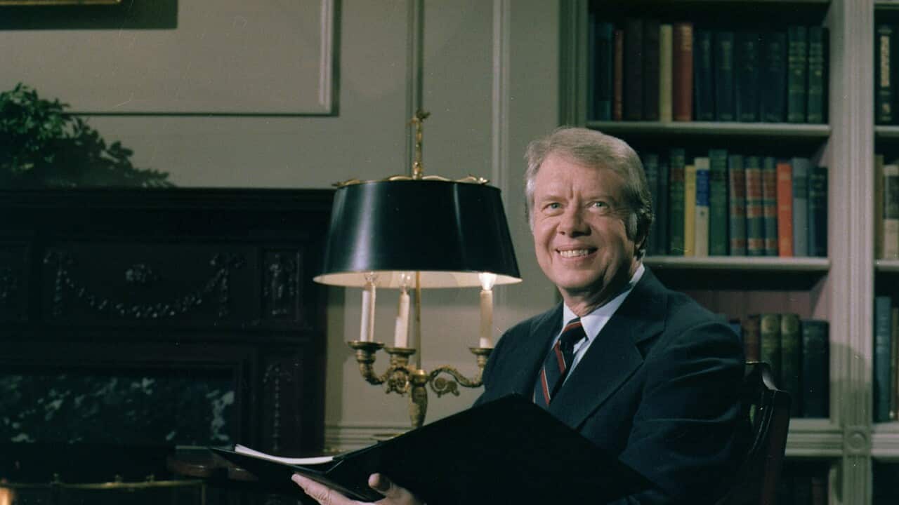 A man wearing a suit and tie smiles as he sits and holds a file.