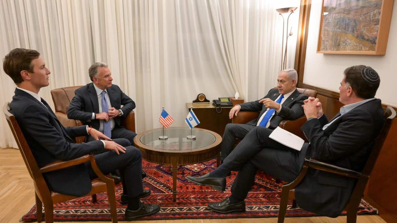 An indoor meeting with four men in suits seated around a small coffee table, with Israeli and US flags displayed on the table.