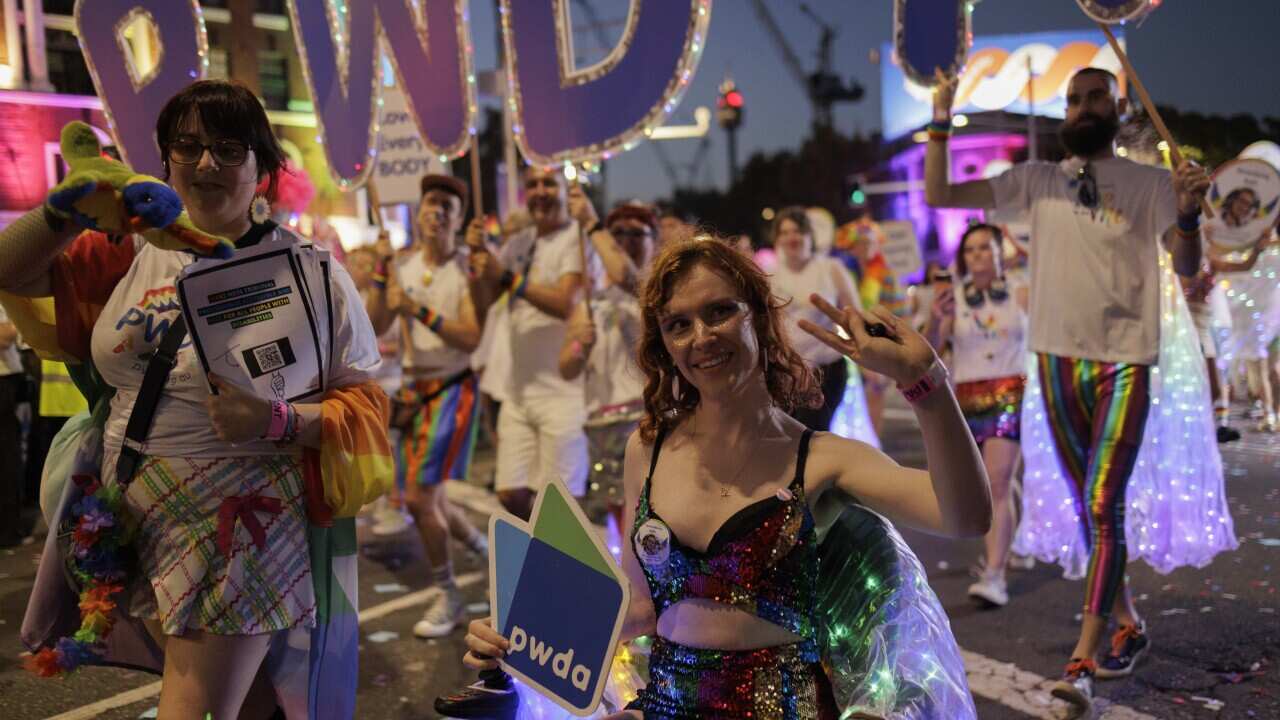 A woman poses for a photo while taking part in Mardi Gras. She is a wheelchair user, is wearing a rainbow sequin top and hotpants, and holding a People With Disbaility Australia sign