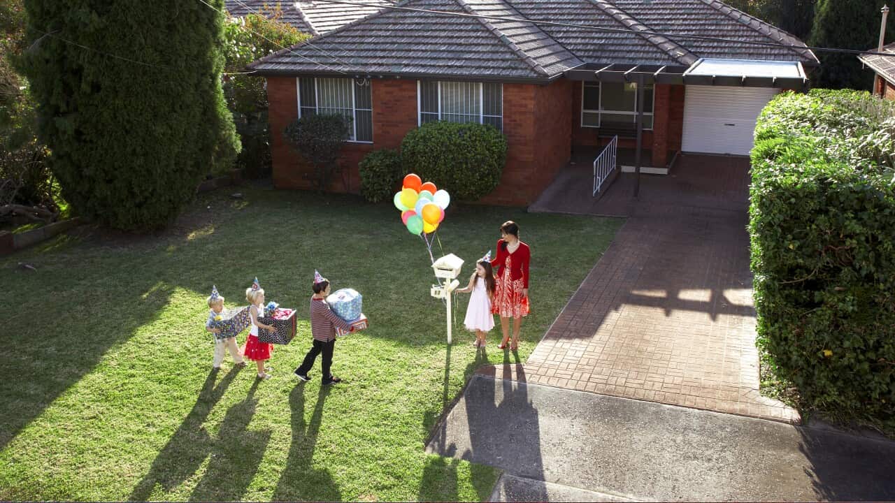 Children holding large presents walking up the front lawn of a house towards a letterbox with balloons on it and a girl with her mum waiting for the guests.