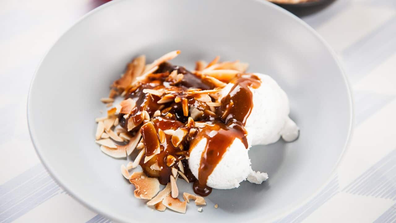 Two scoops of ice-cream sit in a bowl, along with banana, coconut and slivered almonds, all drizzled with a dark caramel sauce.
