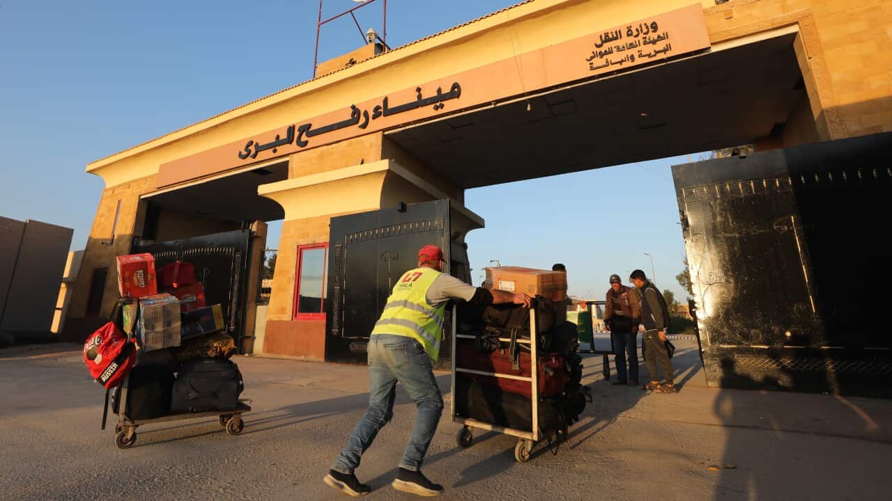 People push carts loaded with luggage towards a border crossing with Arabic writing emblazoned on it.