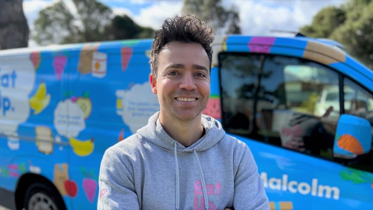A man in a grey hoodie smiles at camera standing next to a colourful van.