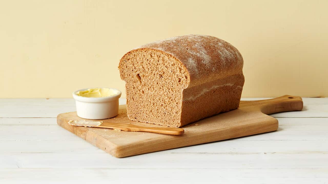 A tall loaf of bread with a wider, rounded top and the front sliced off sits on a wooden board. A pot of butter and a knife sit in front of the bread, on the board.