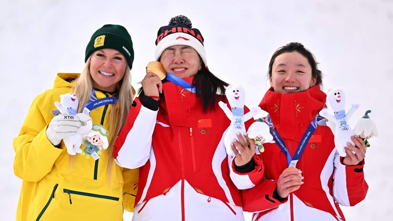 Three female Olympic medalists in winter gear smile on a podium, holding their medals and mascots.