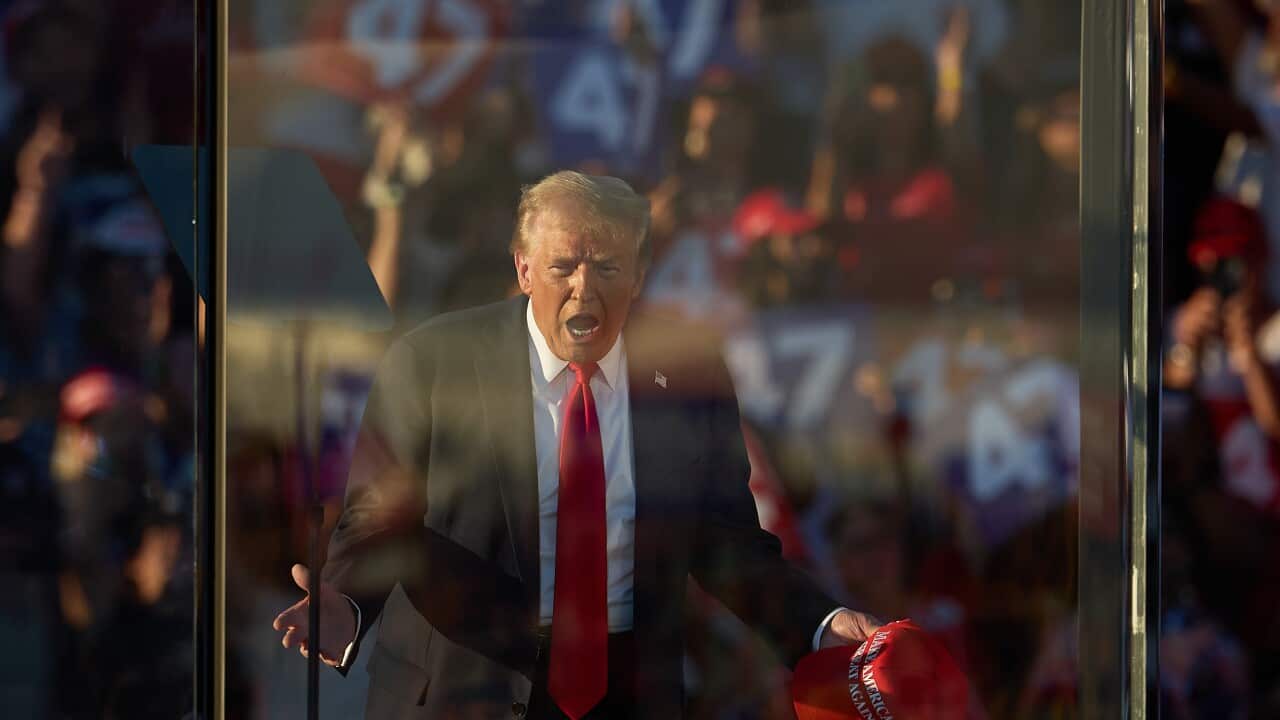 Donald Trump, in a suit and a tie, gestures behind a bulletproof glass.