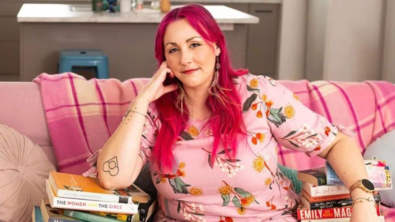 Bigambul and Wakka Wakka author Melanie Saward has bright pink hair and dark brown eyes. She sits on a pink lounge surrounded by stacks of books, looking into the camera.
