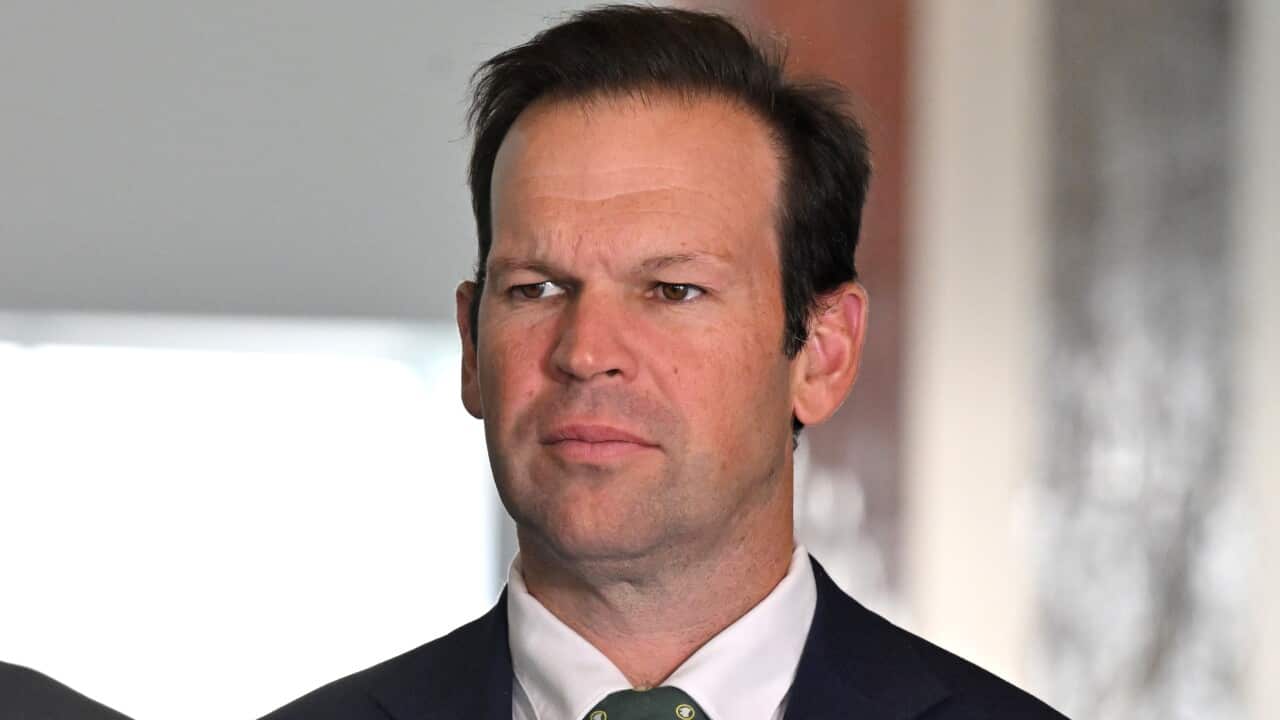A man in a black suit and tie with a stern expression.