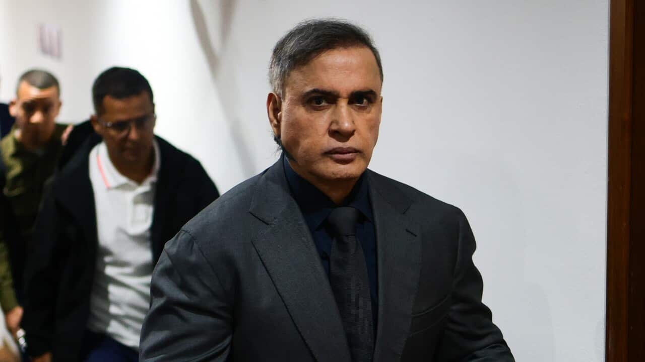 A man in a dark suit and tie holding a blue folder walks through a crowded indoor space while several people follow behind him.