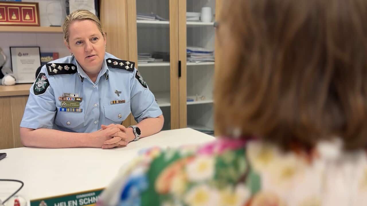 A female police officer in uniform speaks to another woman sitting across from her at a desk.