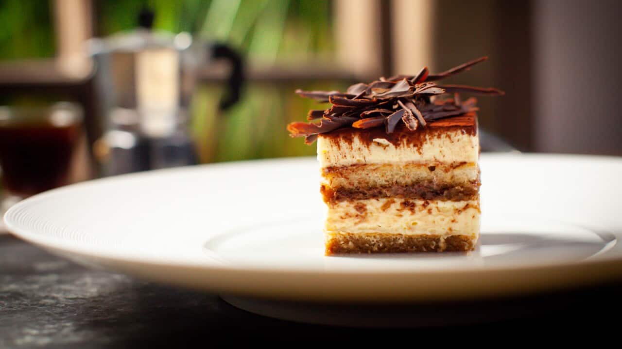 A slice of tinramisue, seen from the side, sits on a wide white plate. The tiramisu piece is topped with shards of chocolate.