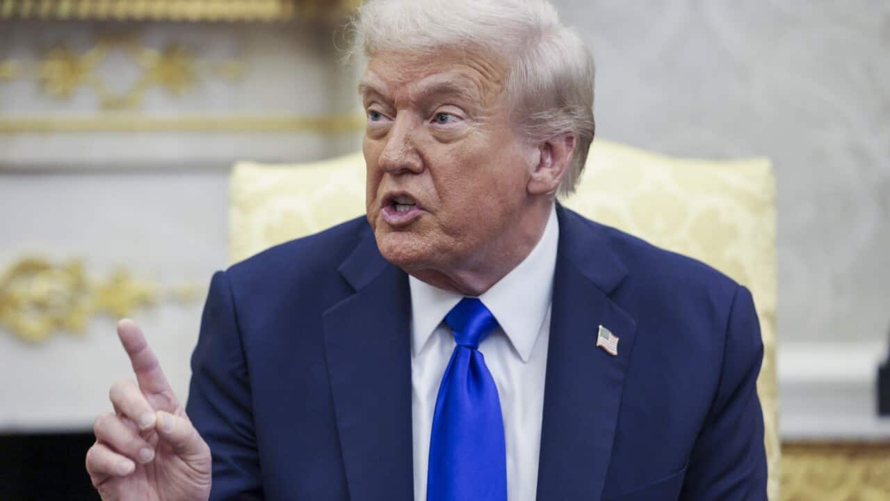 Donald Trump, wearing a dark blue suit and a bright blue tie, is pictured from the chest up, gesturing with an upraised index finger as he speaks. A gold-trimmed fireplace and a yellow armchair are visible behind him.