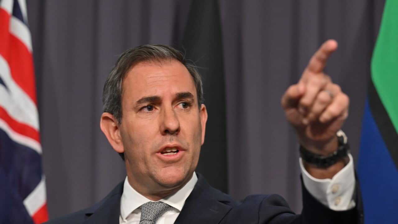 A man in a dark suit with a red tie, with the Australian and Aboriginal flags visible in the background.