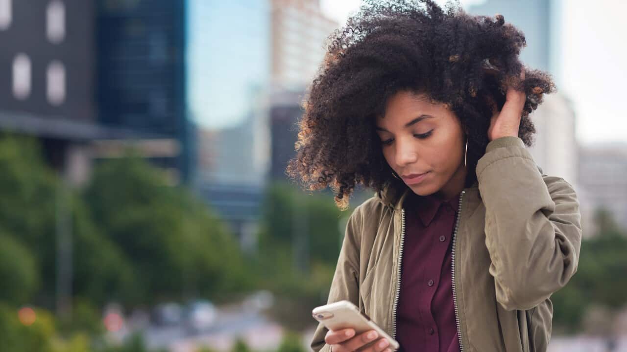 A woman with short, curly hair looks at her phone while buildings are visible behind her.