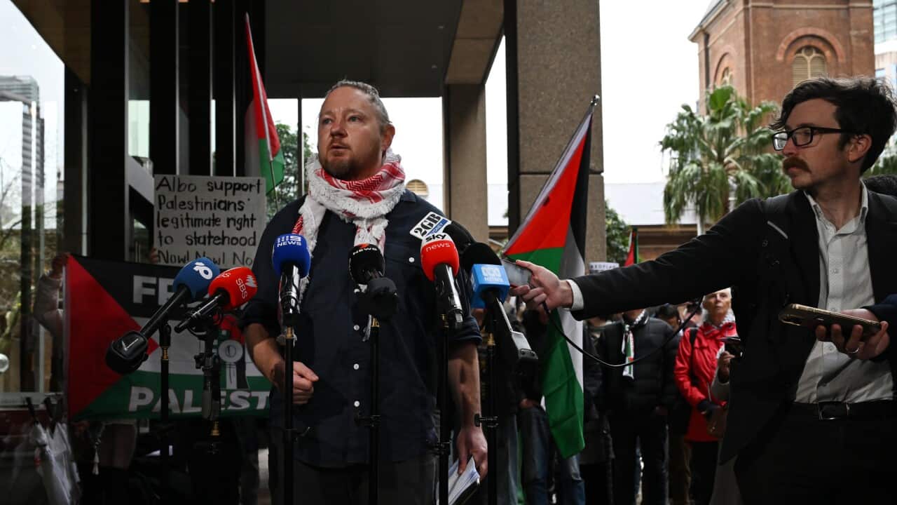A white man wearing a scarf speaking at a press conference outside the NSW Supreme Court. Behind him, protesters hold placards and Palestinian flags.